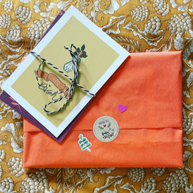 Ein in apricotfarbenes Seidenpapier gepacktes Geschenk, zugeklebt mit einem Fräulein Weichmut Sticker und zwei zusammengebundene Blumen-Postkarten.